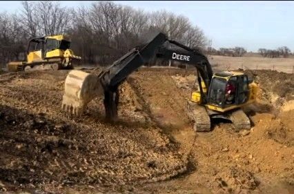 Two excavators working in a muddy dirt lot, one yellow machine in the background and a black Deere in front.
