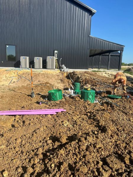 Construction site beside black metal building, with dirt trench, green buckets, pipes, and a worker kneeling.