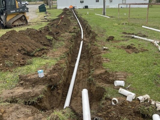 Trenched yard with a long white pipe laid beside a house and excavator in the background
