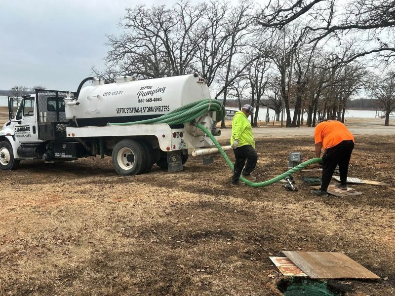 Two workers operate a green hose connected to a white septic pump truck in a field near a lake.