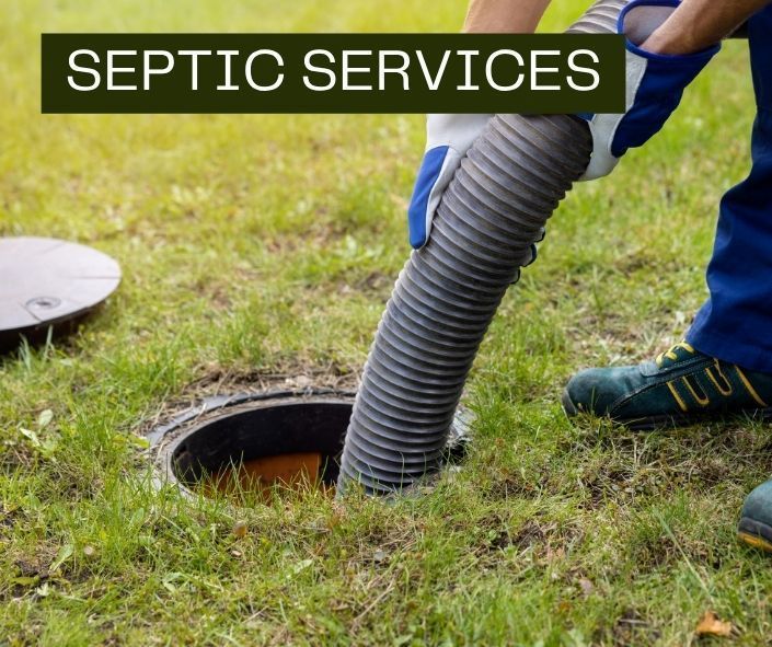 A worker in blue clothing uses a large vacuum hose to pump out an open residential septic tank in a grassy yard.