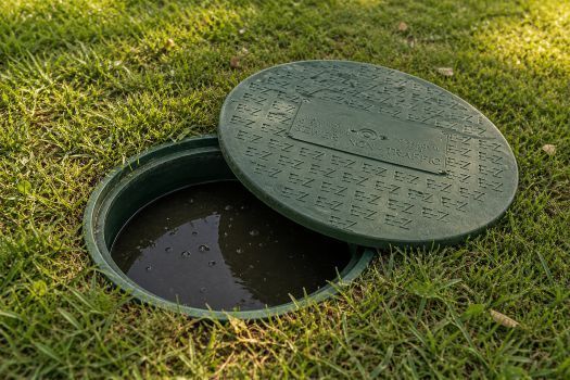 Open green septic cover in grass, revealing a dark full septic tank