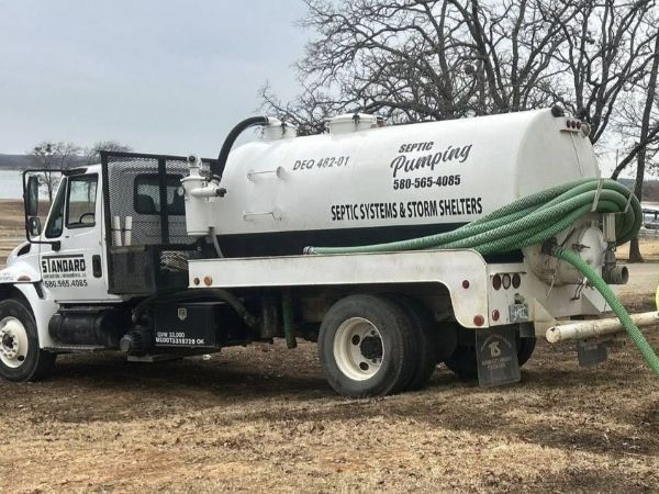 White sewer service truck with a large tank and green hoses parked on grass near bare trees