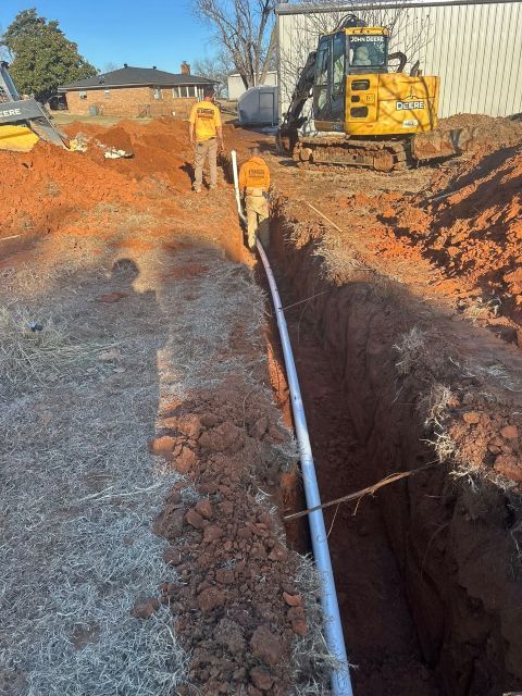 Construction workers installing a white pipe in a deep trench beside a yellow excavator at a worksite