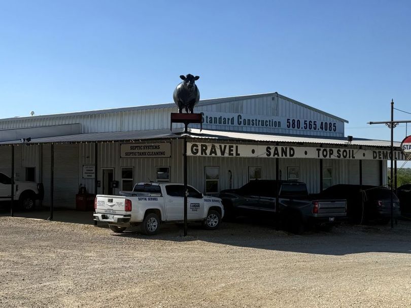 Rural septic, gravel sand, and topsoil store with parked trucks and a large bull statue on the roof under a clear blue sky