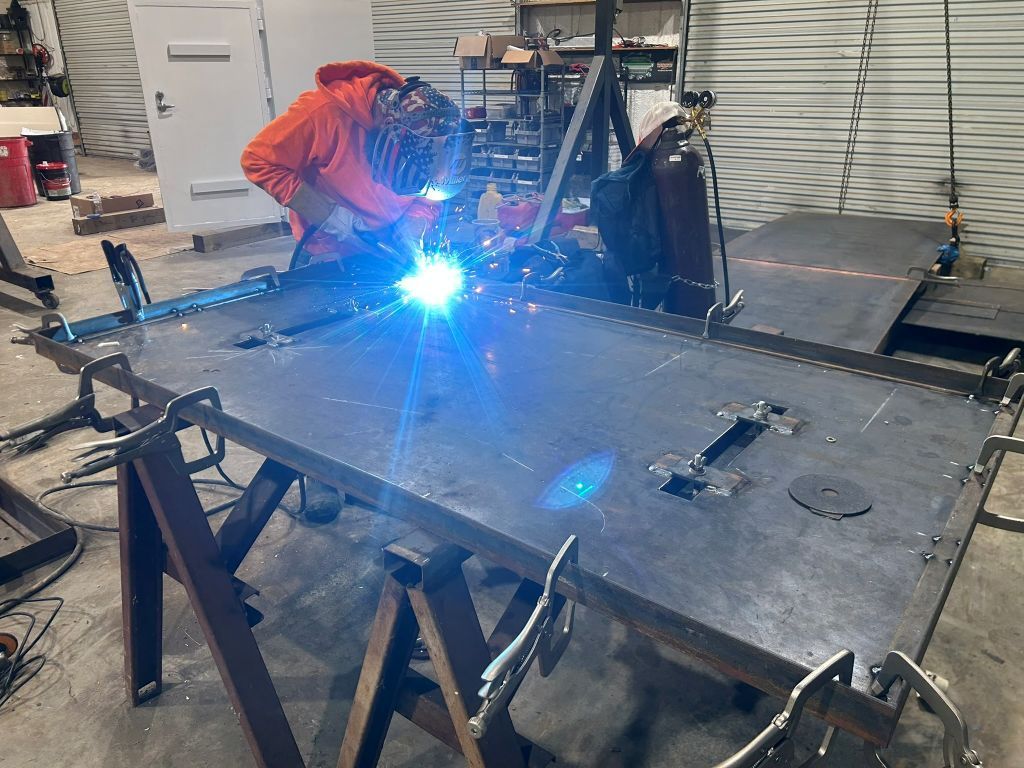 Welder in orange jacket welding a large metal sheet on a workbench, bright blue arc in a workshop