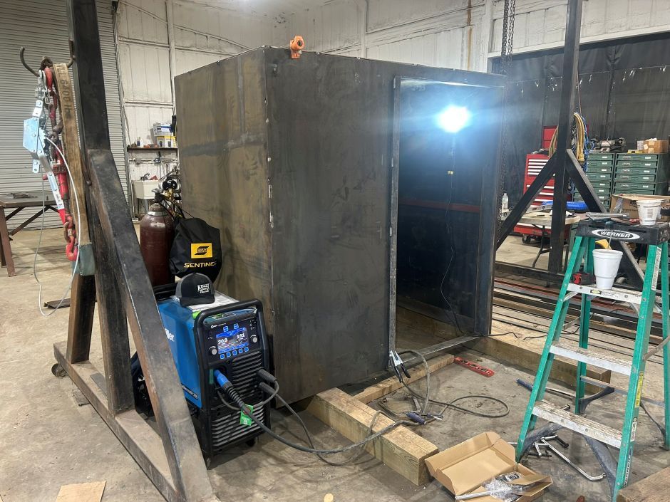 Large black metal enclosure in a workshop, with a bright light inside and tools, ladder, and generator nearby.