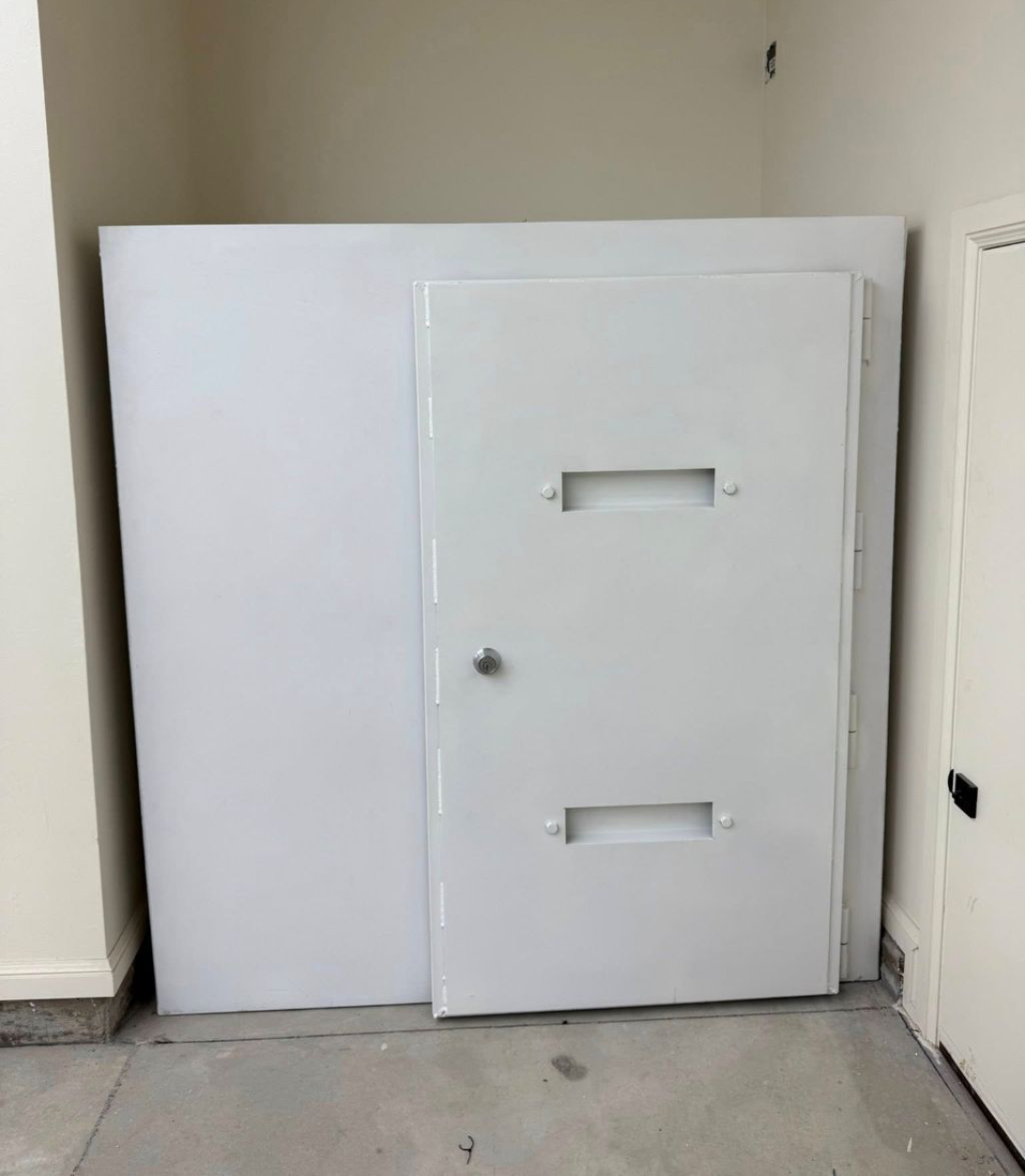 White door on an indoor storm shelter safe room in a bare indoor hallway, resembling a secure access door