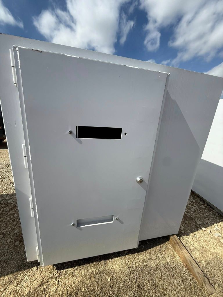Gray metal tonado shelter with two vents and a slanted top beside a white wall outdoors under a blue sky.