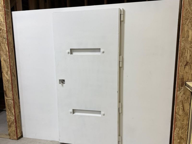 Large white steel safe room storm shelter with two horizontal handles in a bare room under construction