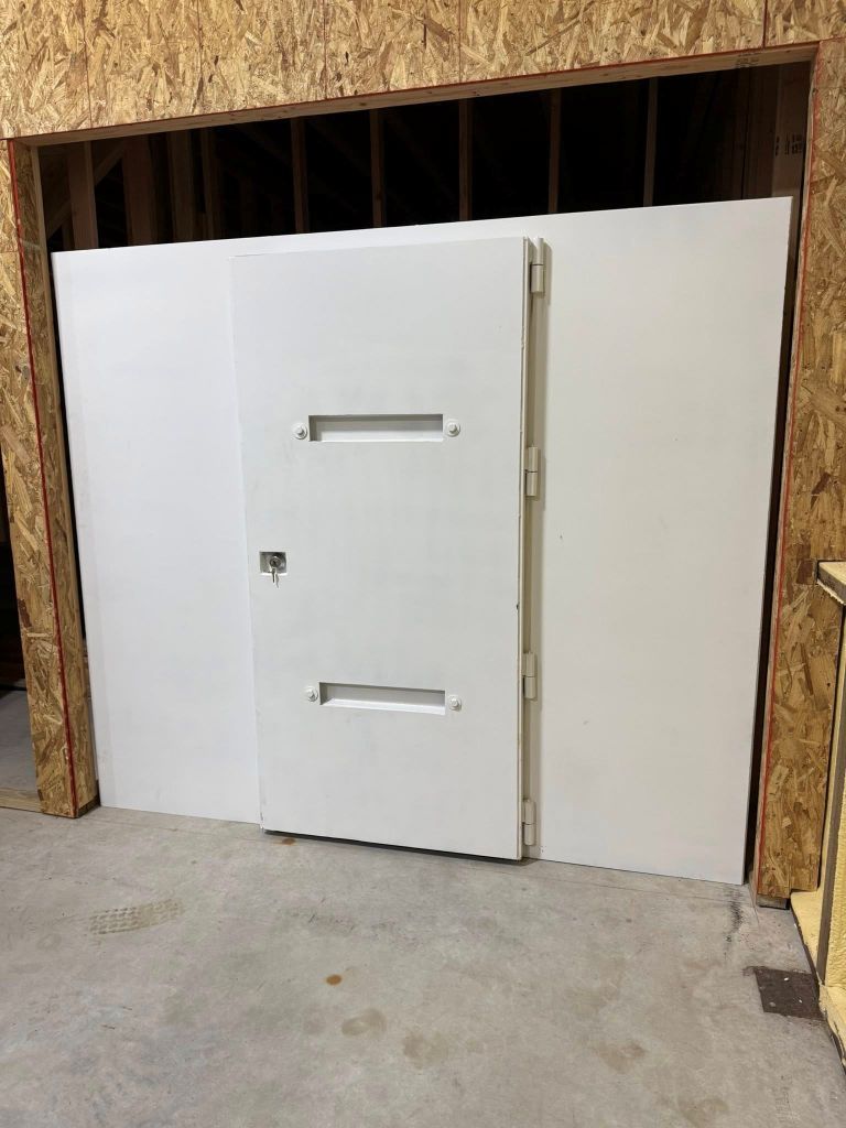 white metal indoor storm shelter with door set in a plywood-walled opening