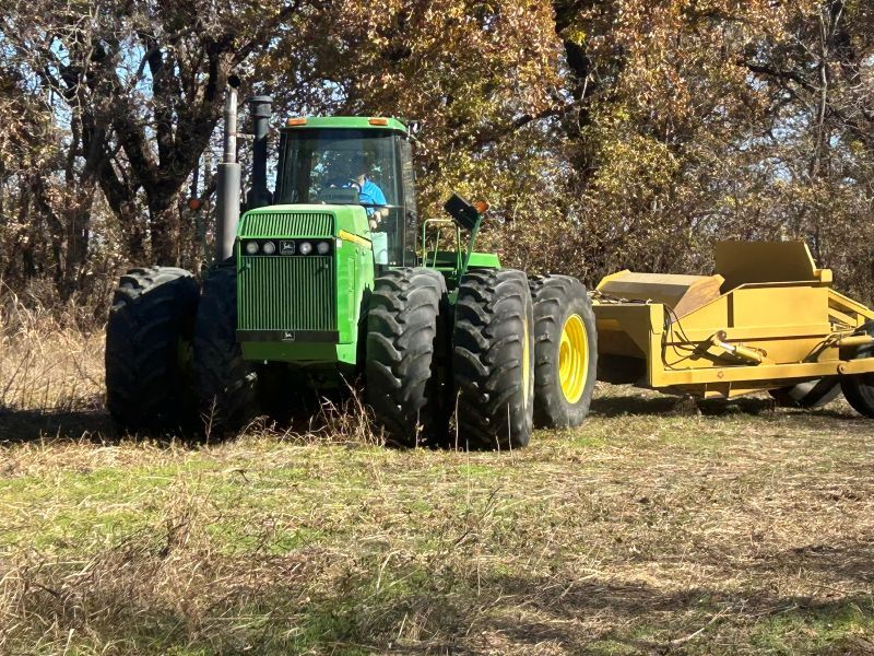 Green tractor with large tires pulling yellow landmover in a field under trees.