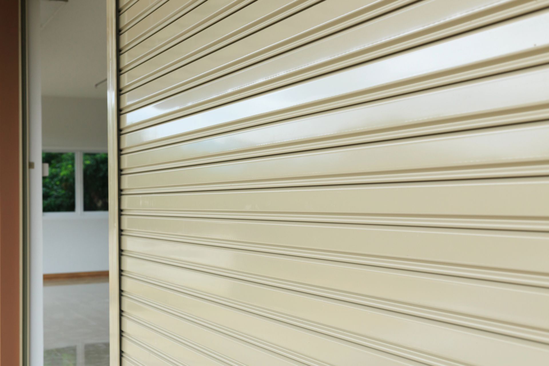 Una serranda avvolgibile in metallo beige chiusa accanto a una porta, con uno scorcio di una stanza interna.