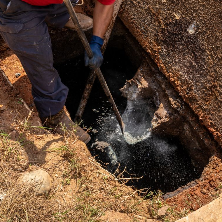 Worker in blue gloves probes a muddy pit with a tool as water splashes up from below.