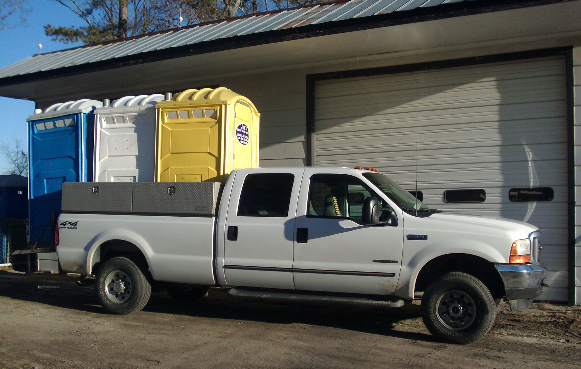 A white truck is parked in front of a garage with two portable toilets in the bed