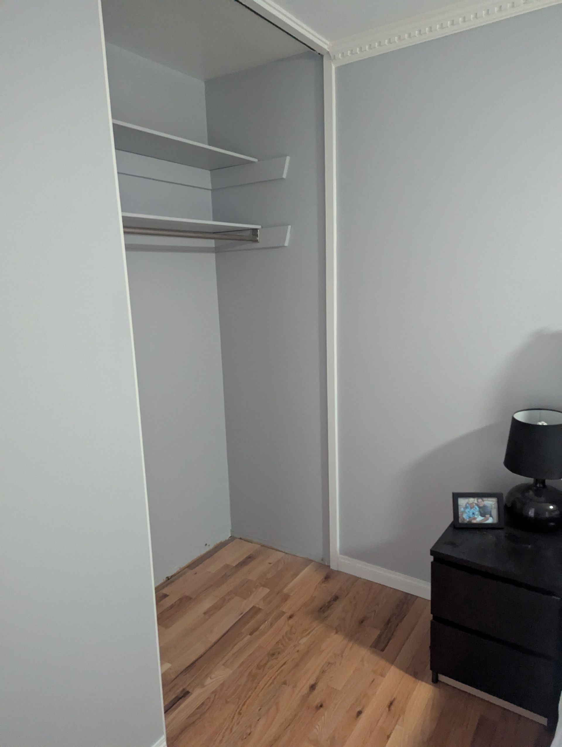 Empty closet with white walls, shelves, and wood flooring beside a black dresser.