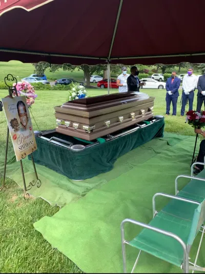 Open casket under a tent at a graveside funeral with mourners nearby
