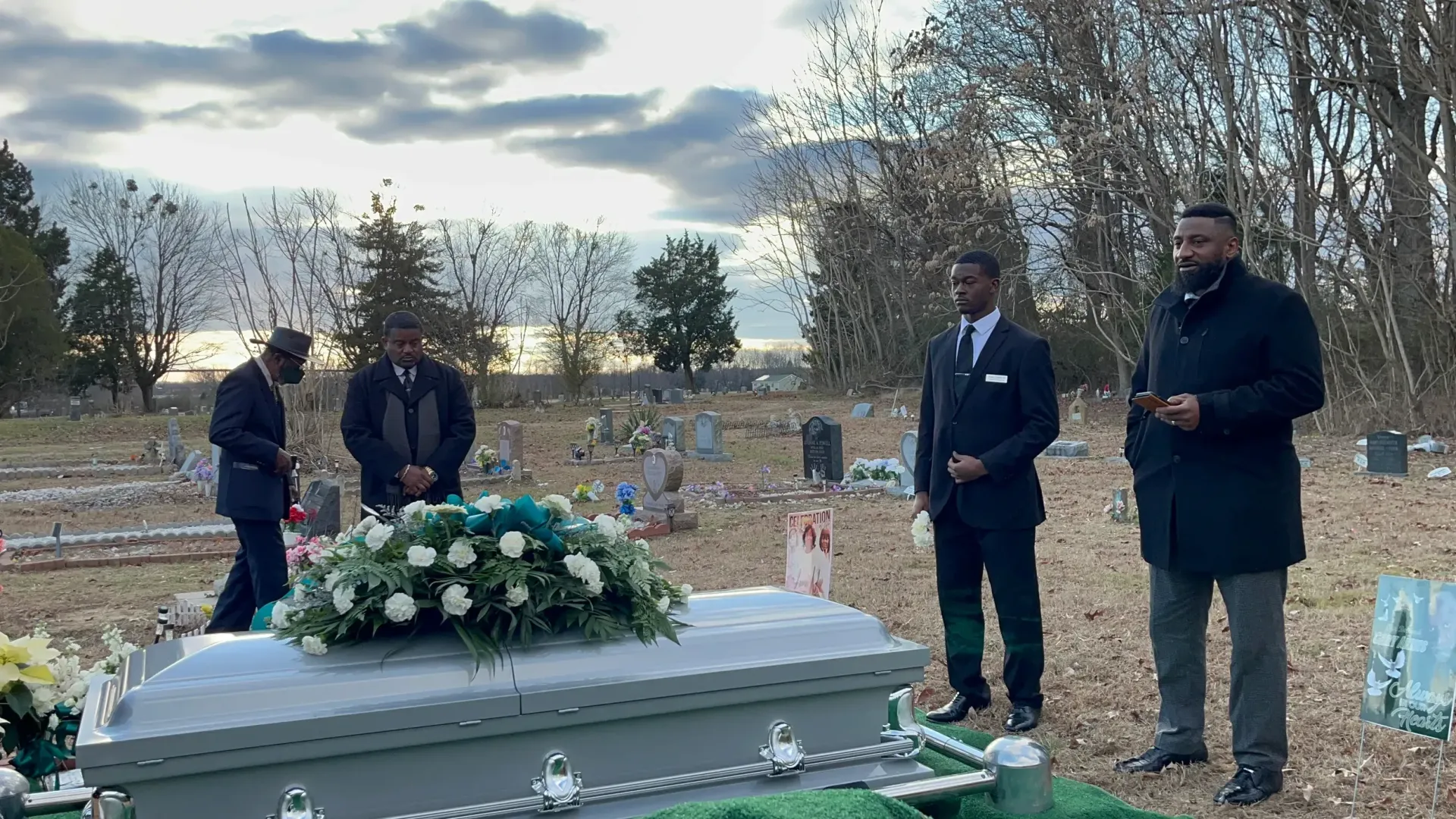 People standing beside a flower-covered casket in an outdoor cemetery at dusk