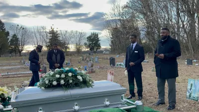 People standing beside a flower-covered casket in an outdoor cemetery at dusk