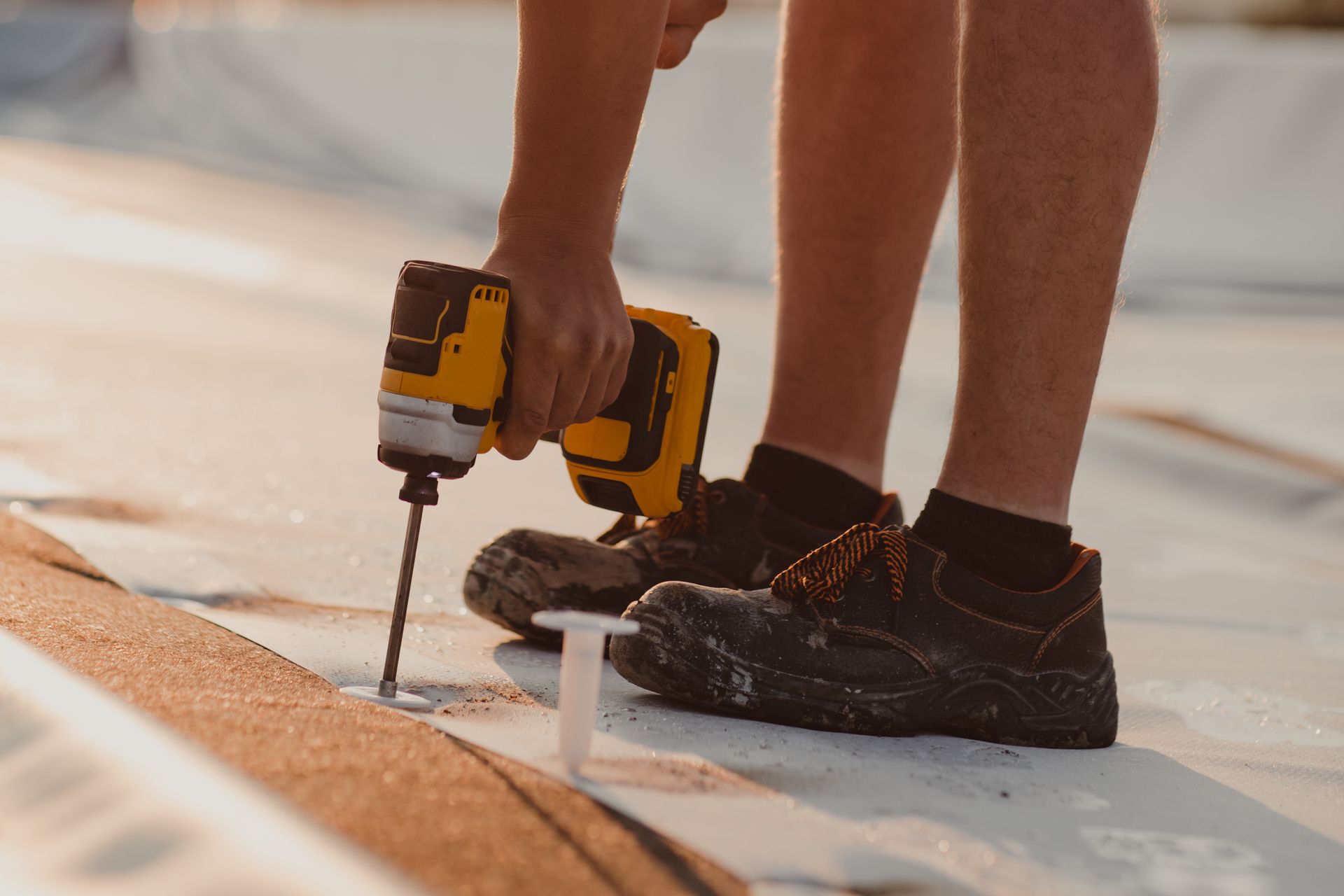 Worker drilling into a white rooftop surface with a yellow power drill and black boots