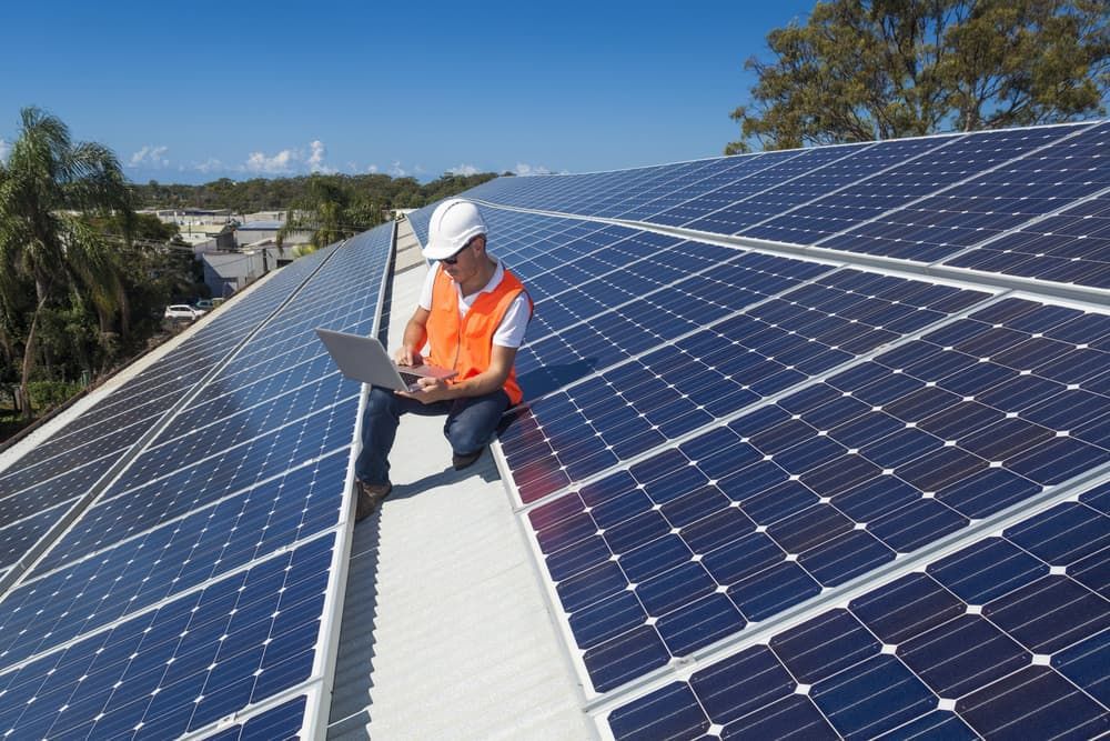 A Man Is Kneeling On Top Of A Roof With Solar Panels And Using A Laptop — Evolution Solar Kingaroy in Goomeri, QLD
