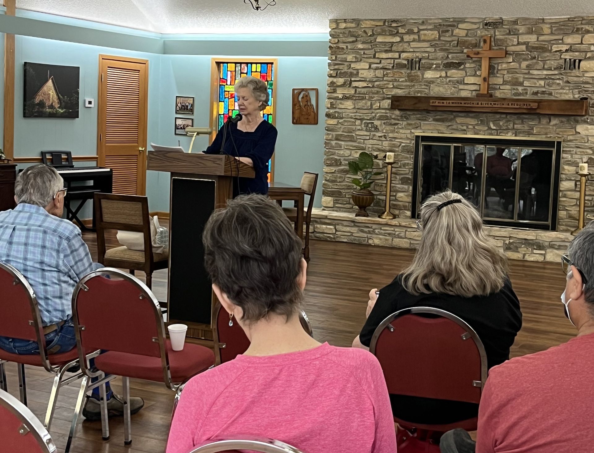 A woman speaks at a podium to an audience in a room with a fireplace.