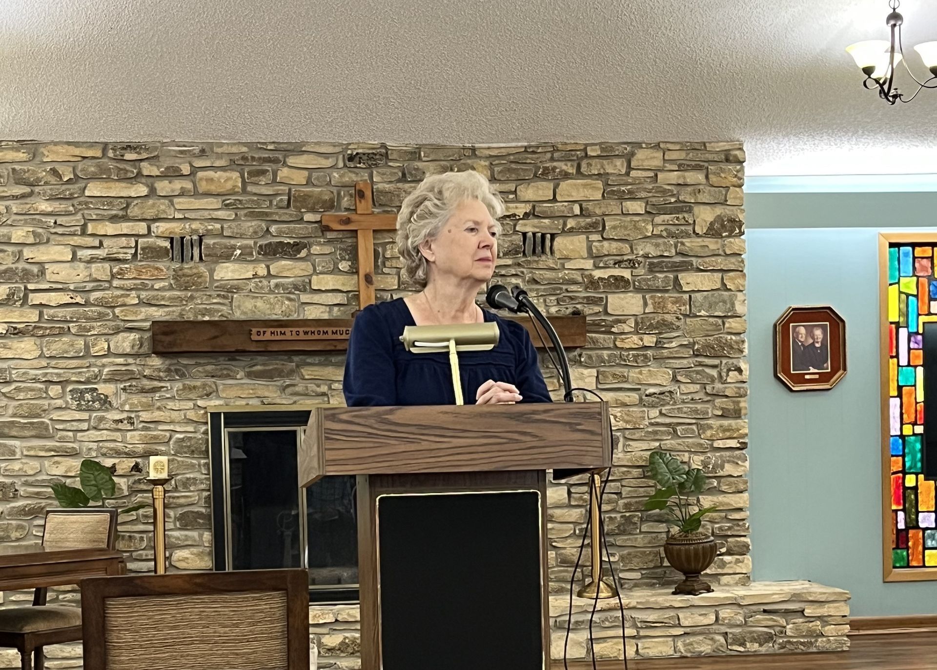 Woman speaking at a podium in a room with a fireplace and a colorful stained-glass window.