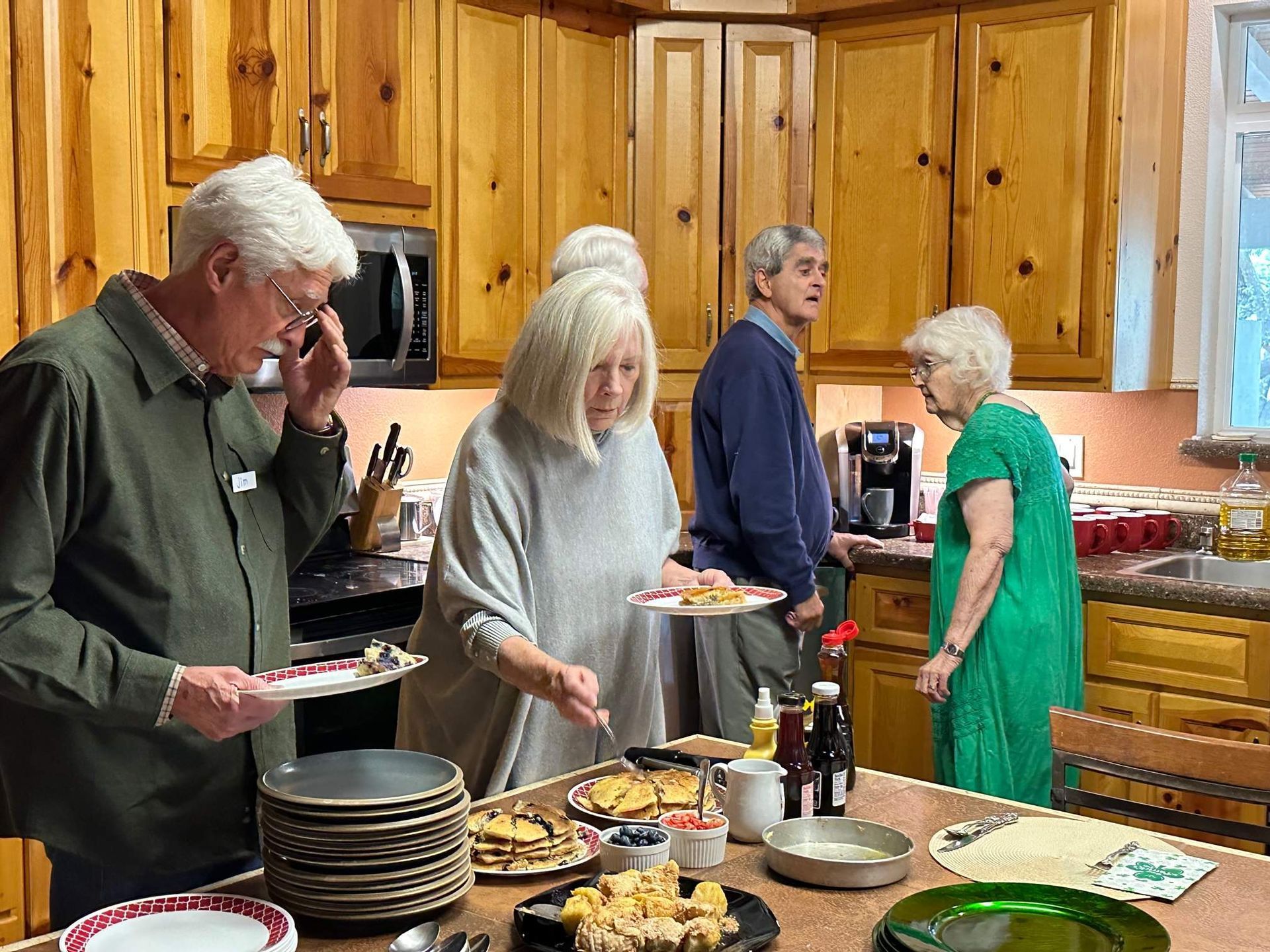 People at a kitchen buffet, serving food. Wooden cabinets, plates, and beverages are visible.