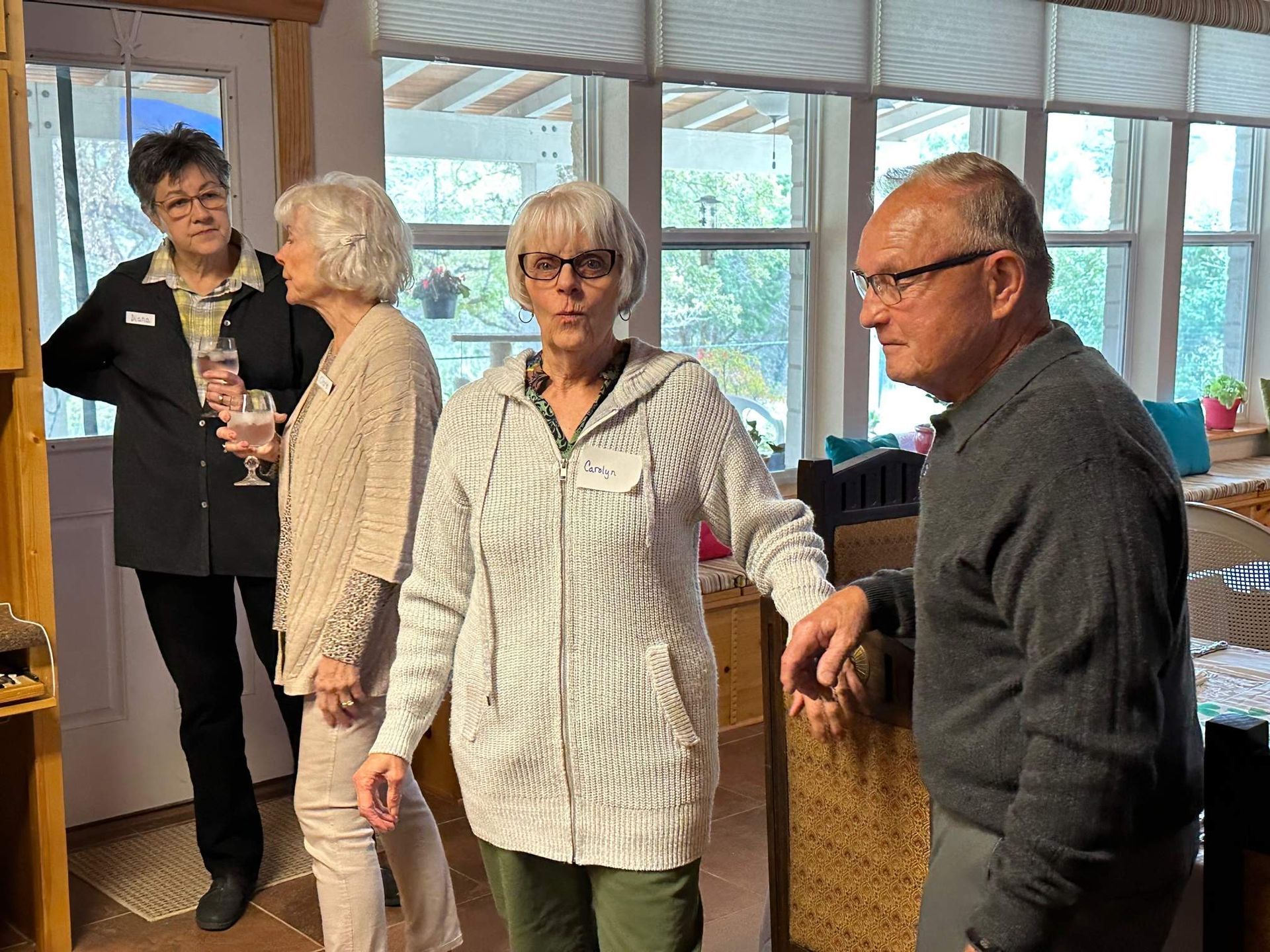 Four people in a room with windows; one in a light gray hoodie, two holding drinks, talking.