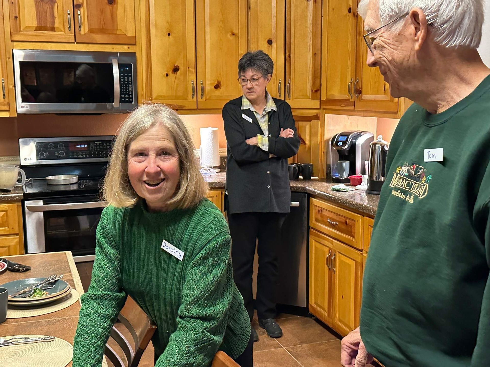 Three people in a kitchen. Woman in green smiles, two others in kitchen: one with arms crossed, another smiles.