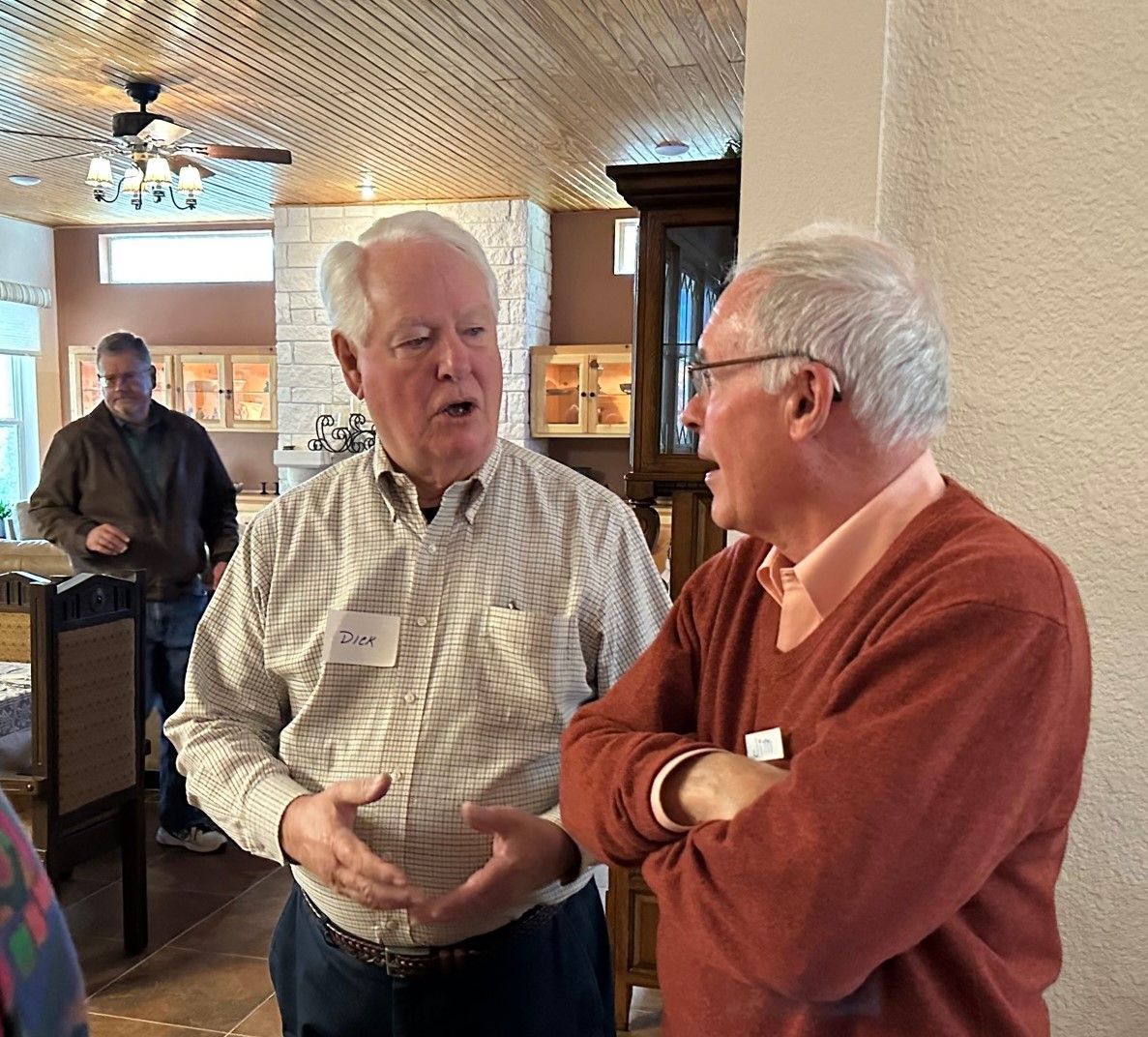 Two men in conversation, one gesturing. Interior setting with other people present.