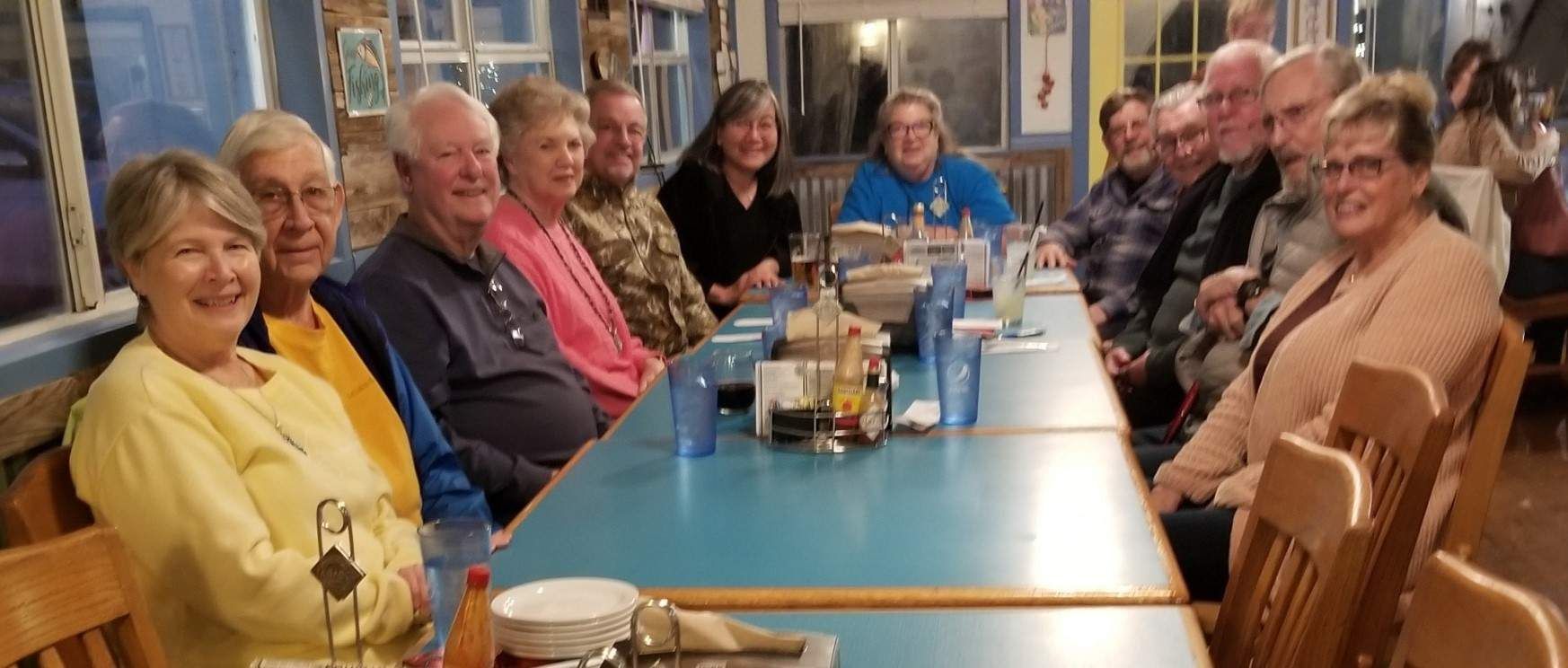 A group of people sitting at a long table in a restaurant, smiling. Blue tabletop, wooden chairs.