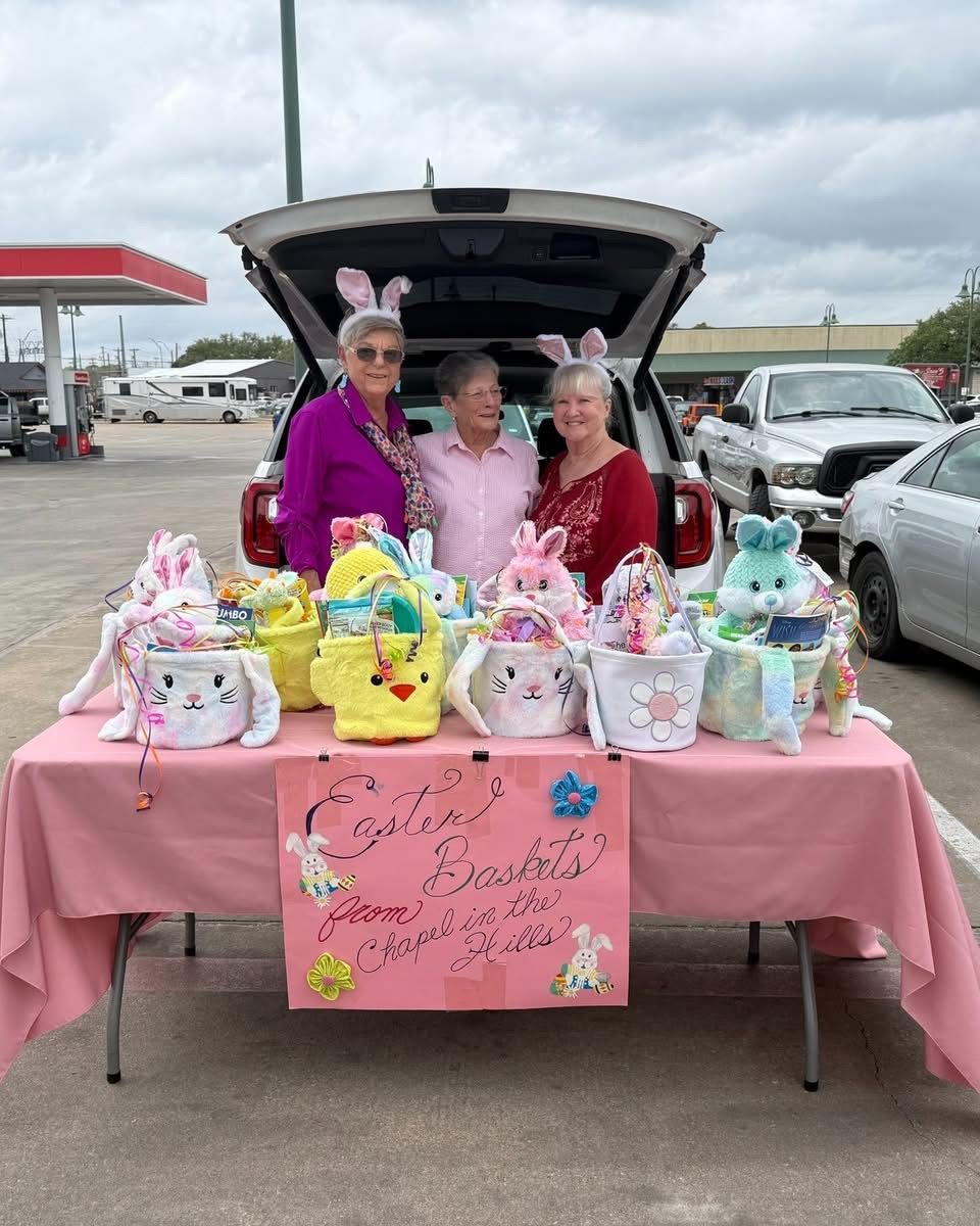 Three people wearing bunny ears stand behind a table filled with Easter baskets in a parking lot.