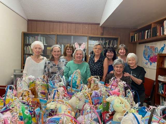 Group of women smiling behind a table piled with Easter baskets in a library setting. One woman wears bunny ears.