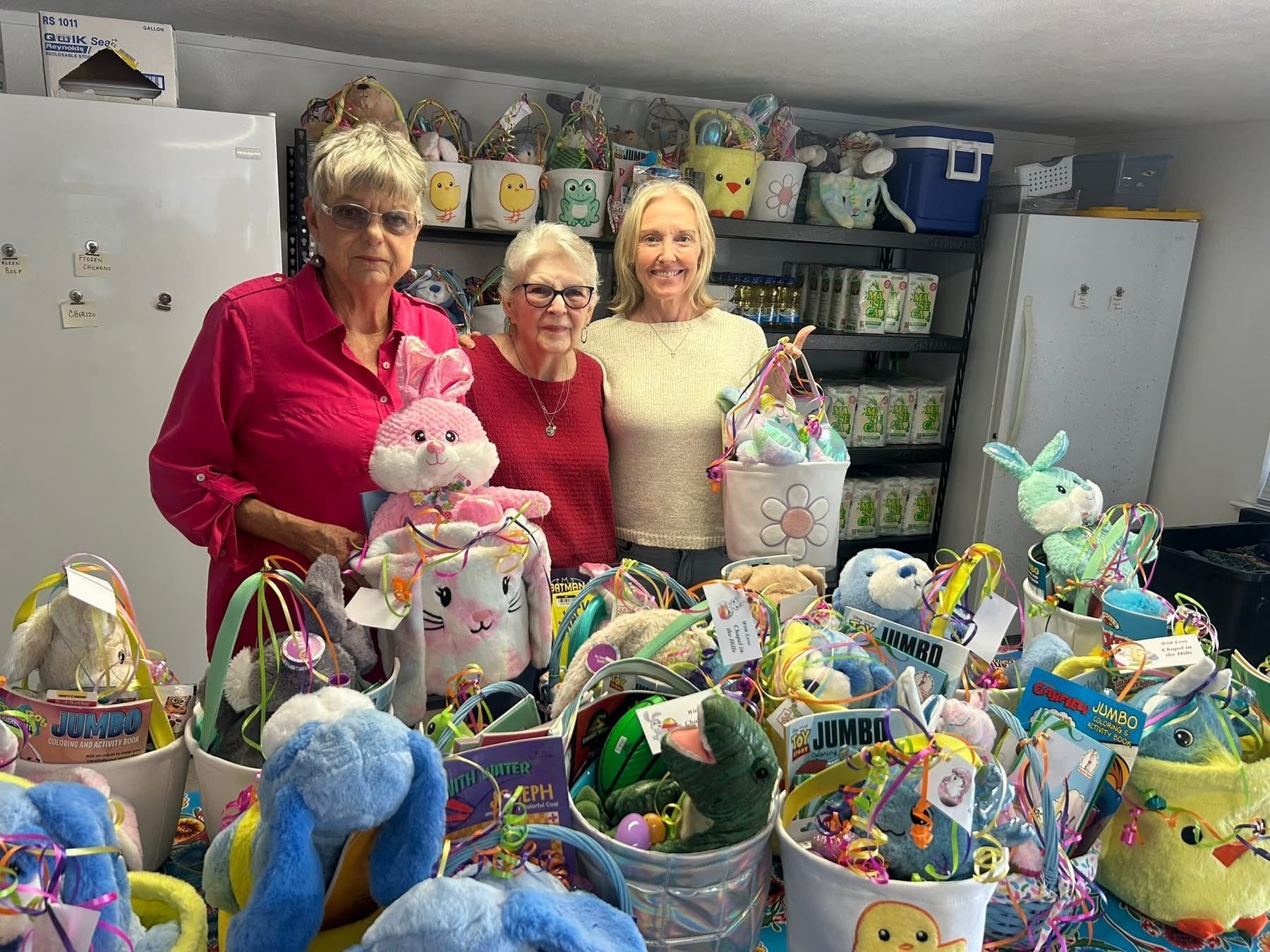 Three people stand behind a table filled with many colorful Easter baskets and plush toys in a storage room.