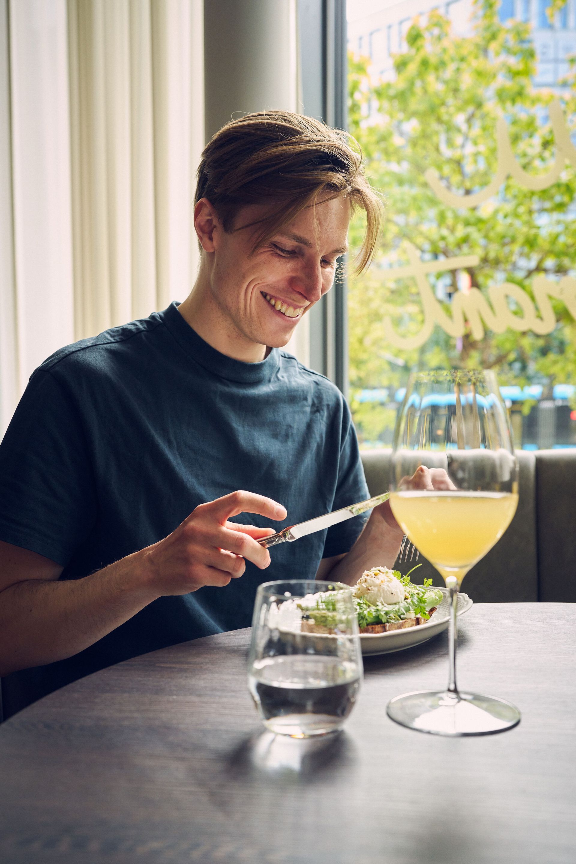 En smilende mann spiser salat med en drink ved et bord. Sollys strømmer inn fra et vindu.