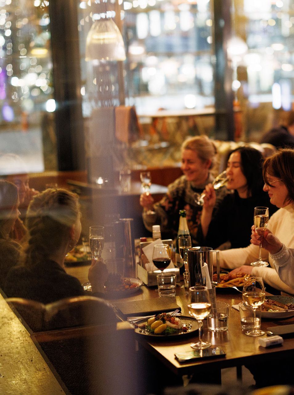 Folk skåler med champagneglass ved et restaurantbord, bylysene er synlige gjennom vinduet.