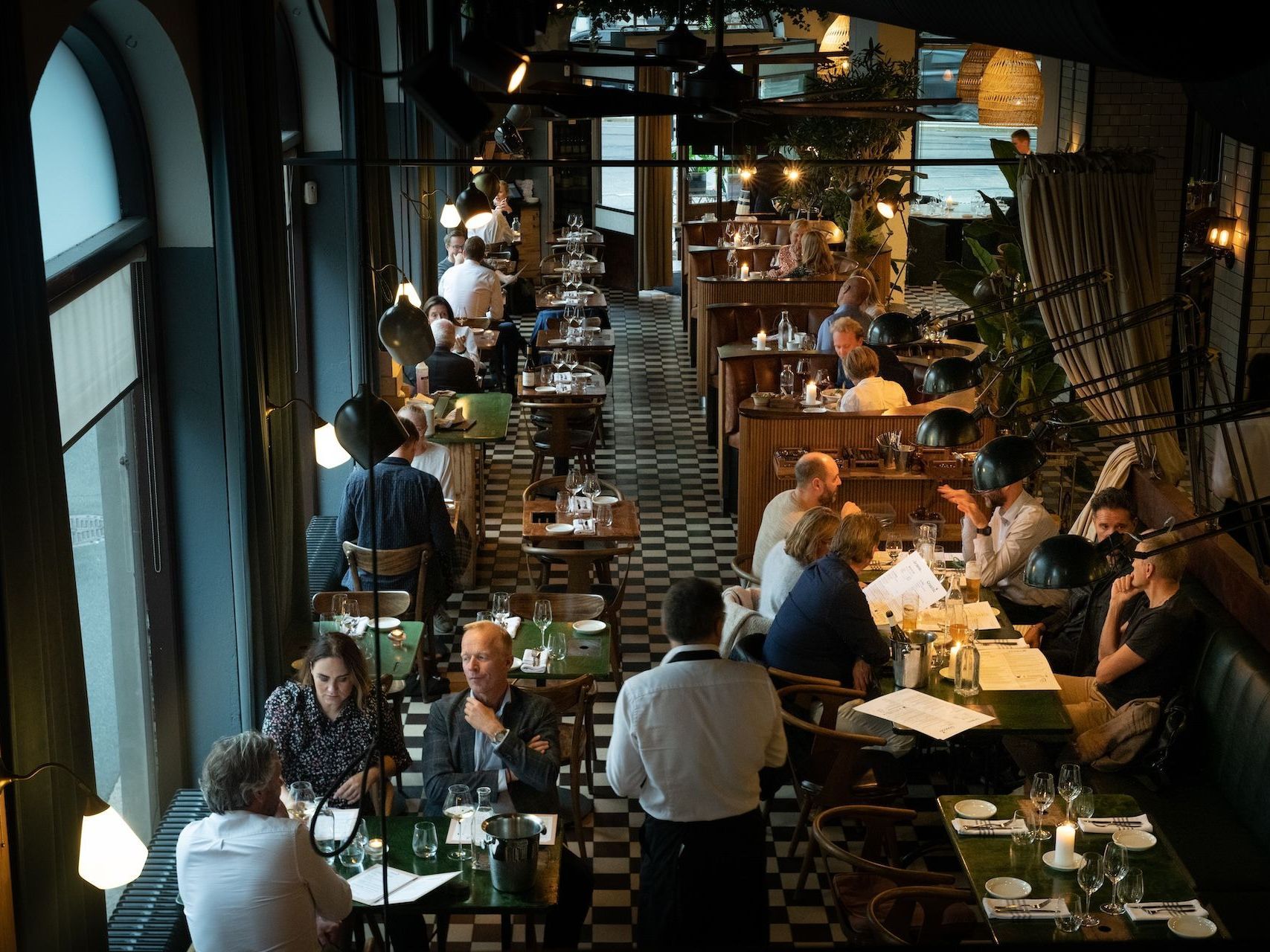 Restaurantinteriør med gjester som spiser, en servitør, svart-hvitt rutete gulv og store vinduer.