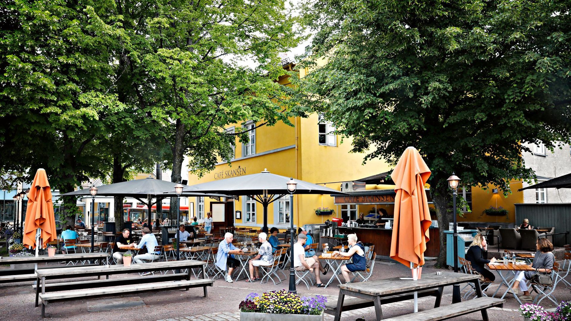 Uteplass med folk som spiser ved bord under parasoller, i nærheten av en gul bygning med trær.