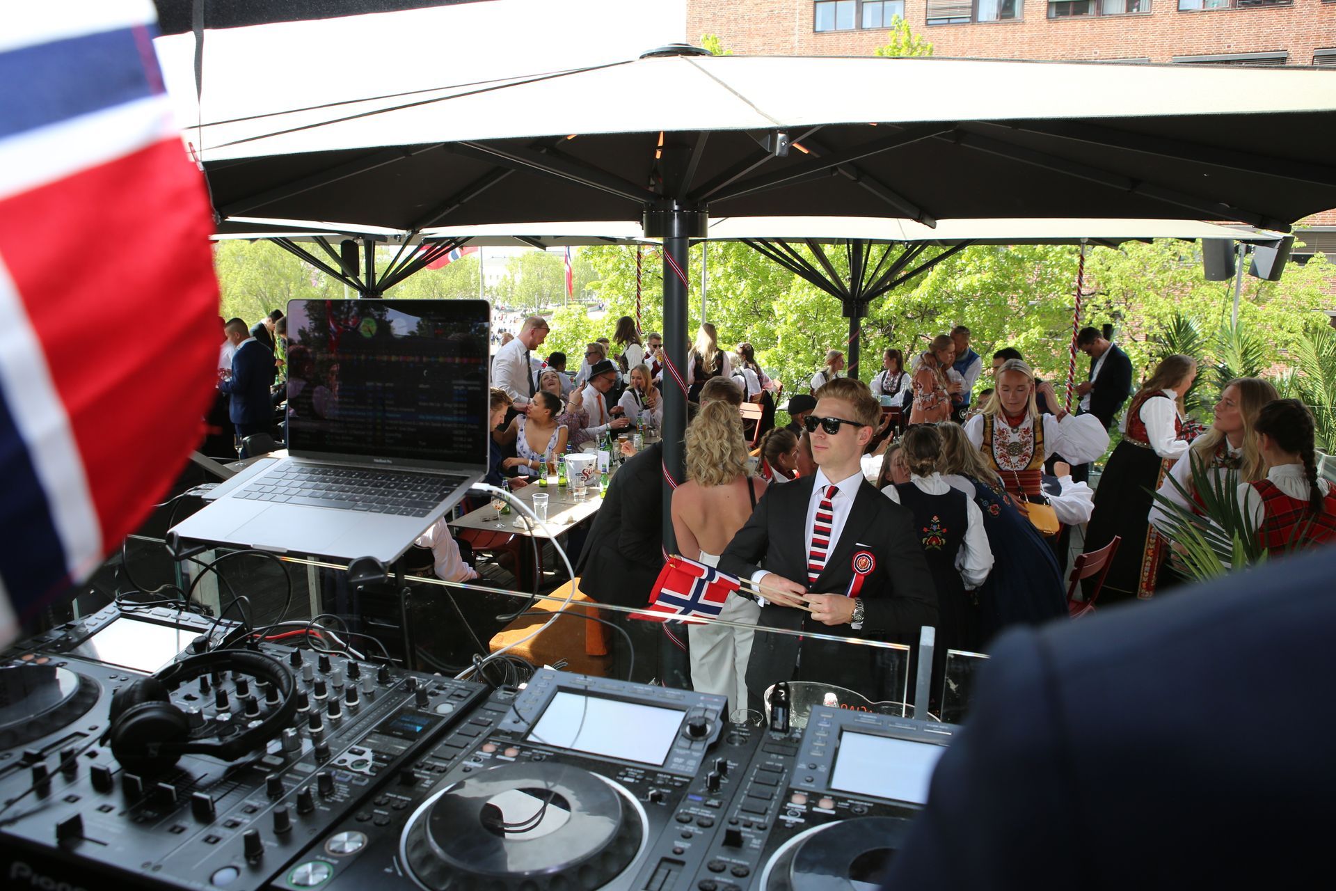 En gruppe mennesker sitter ved bordene under paraplyer mens en dj spiller musikk.