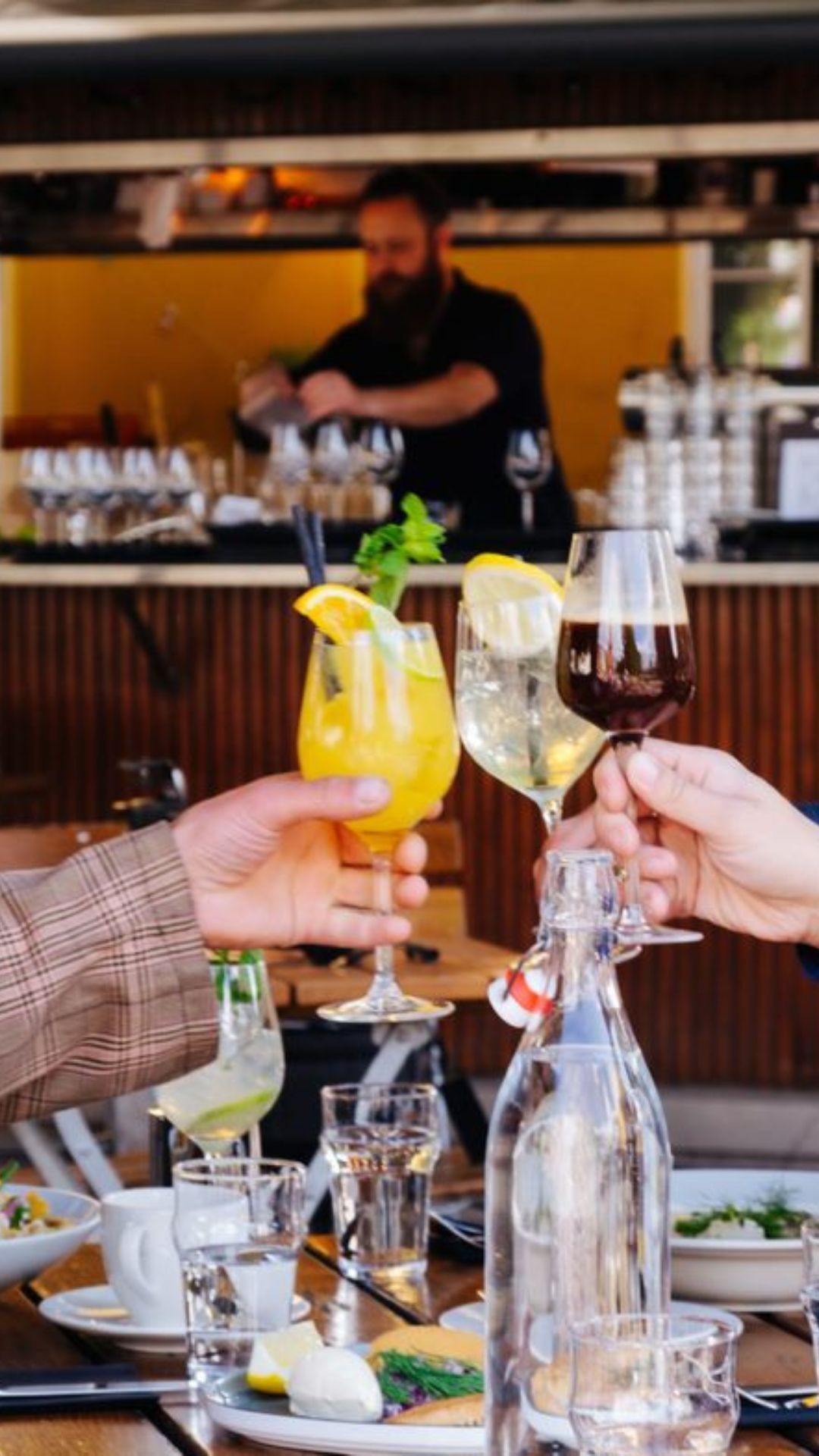 Folk skåler for drinker på en utendørsbar, bartender i bakgrunnen, bord dekket med mat og vannflaske.