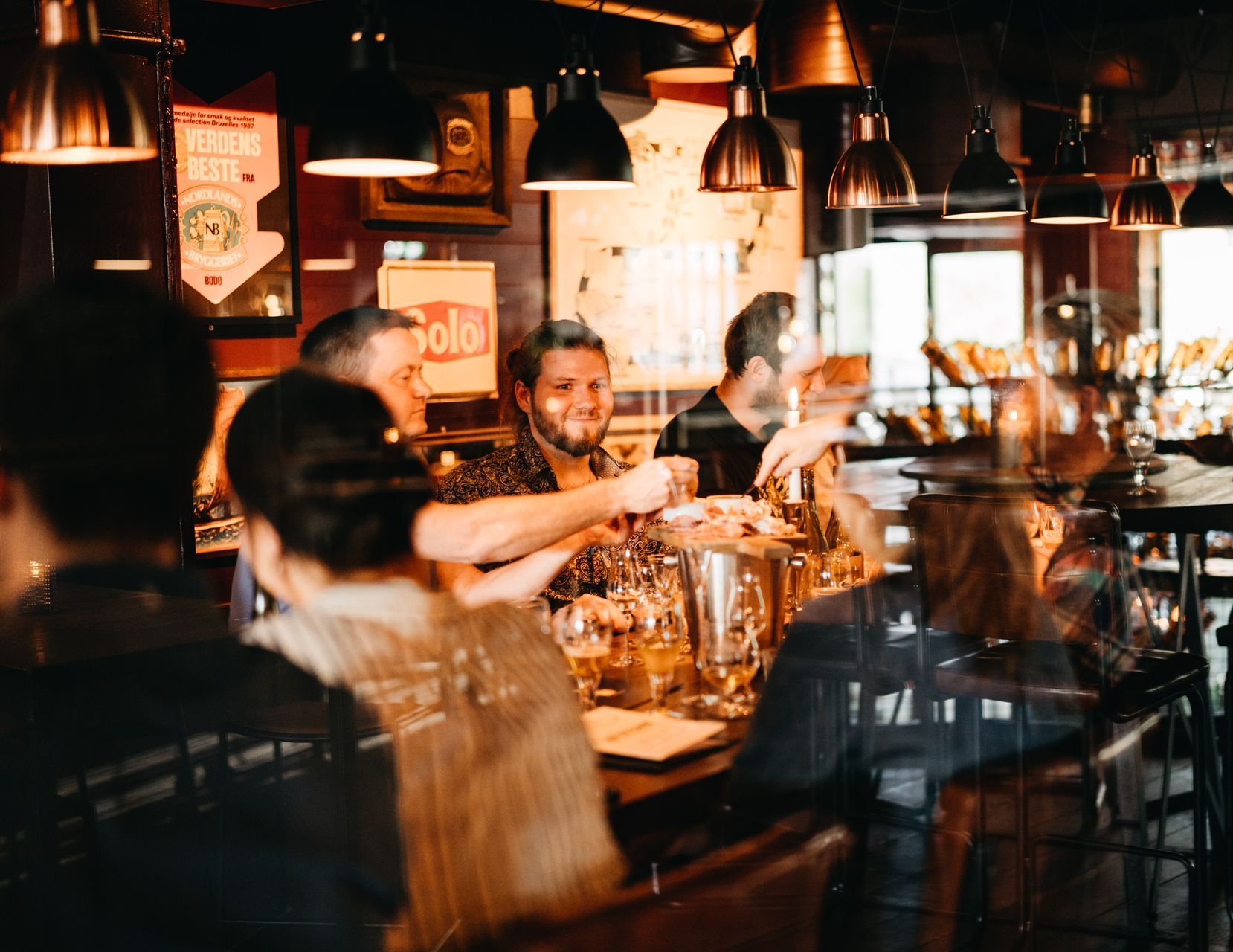 Folk i en bar, noen tar seg av drinker. Dempet belysning, mørkt treverk og hengende lamper.