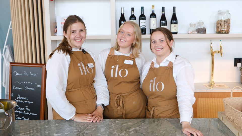 Tre personer i forklær bak en disk, smilende. Butikkinteriør med flasker på en hylle. Forklær har «ilo»-logoen.