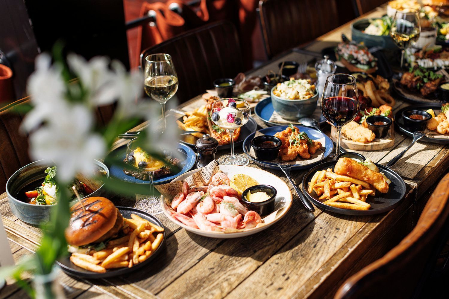 Matbord: burgere, pommes frites, reker, salater, vin og andre retter på et trebord.