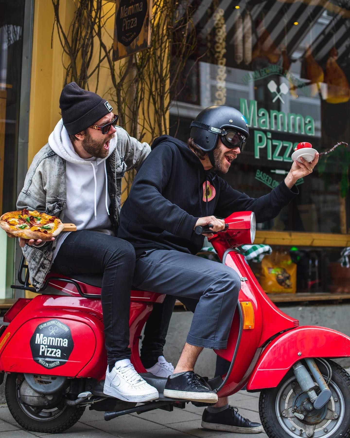 To menn kjører på en rød scooter og en av dem holder en pizza.