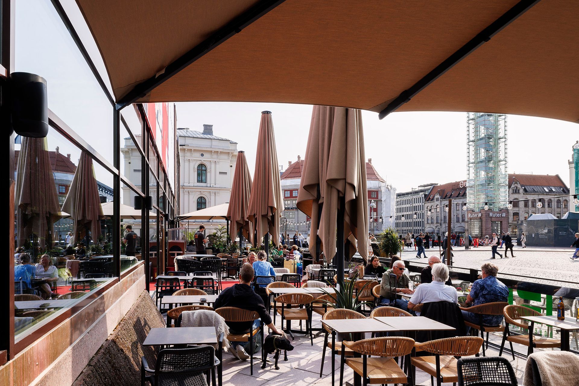 Utendørskafé med gjester, bord og lukkede parasoller på et torg på en solrik dag.
