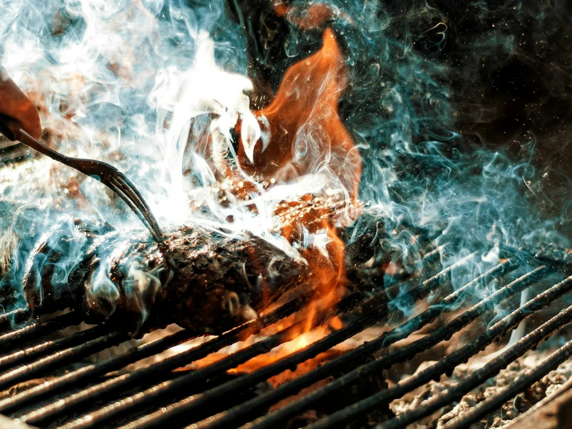 Hamburgergrilling på en røykfylt grill med flammer og en hånd som holder en gaffel.