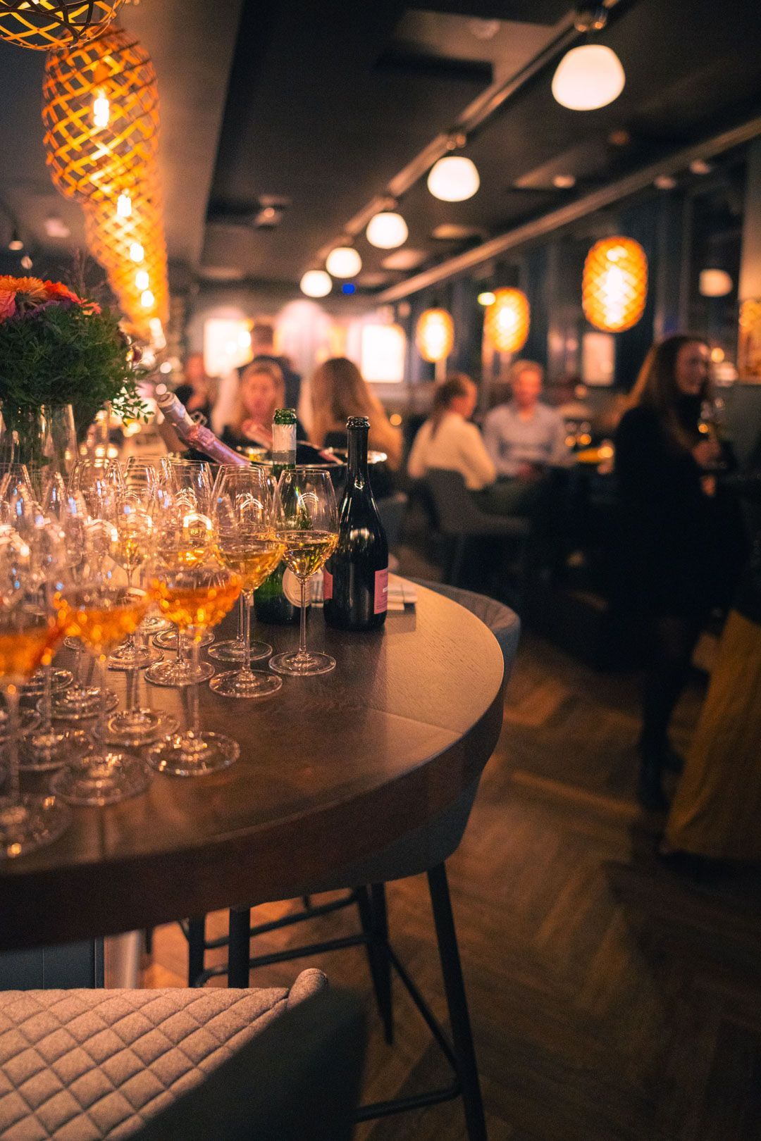 Vinglass på et rundt bord i en bar med sittende folk, opplyst av varme lys i taket.
