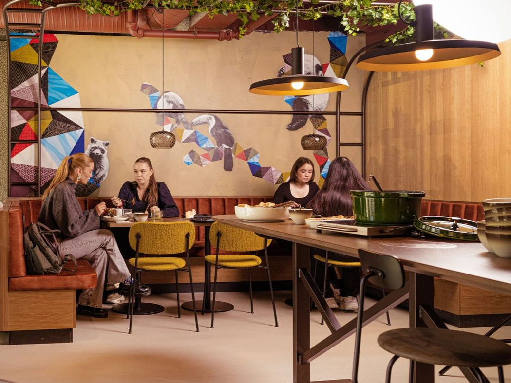 Restaurantinteriør med kunder sittende ved bord; kunst på veggene, gule stoler og pendellamper.