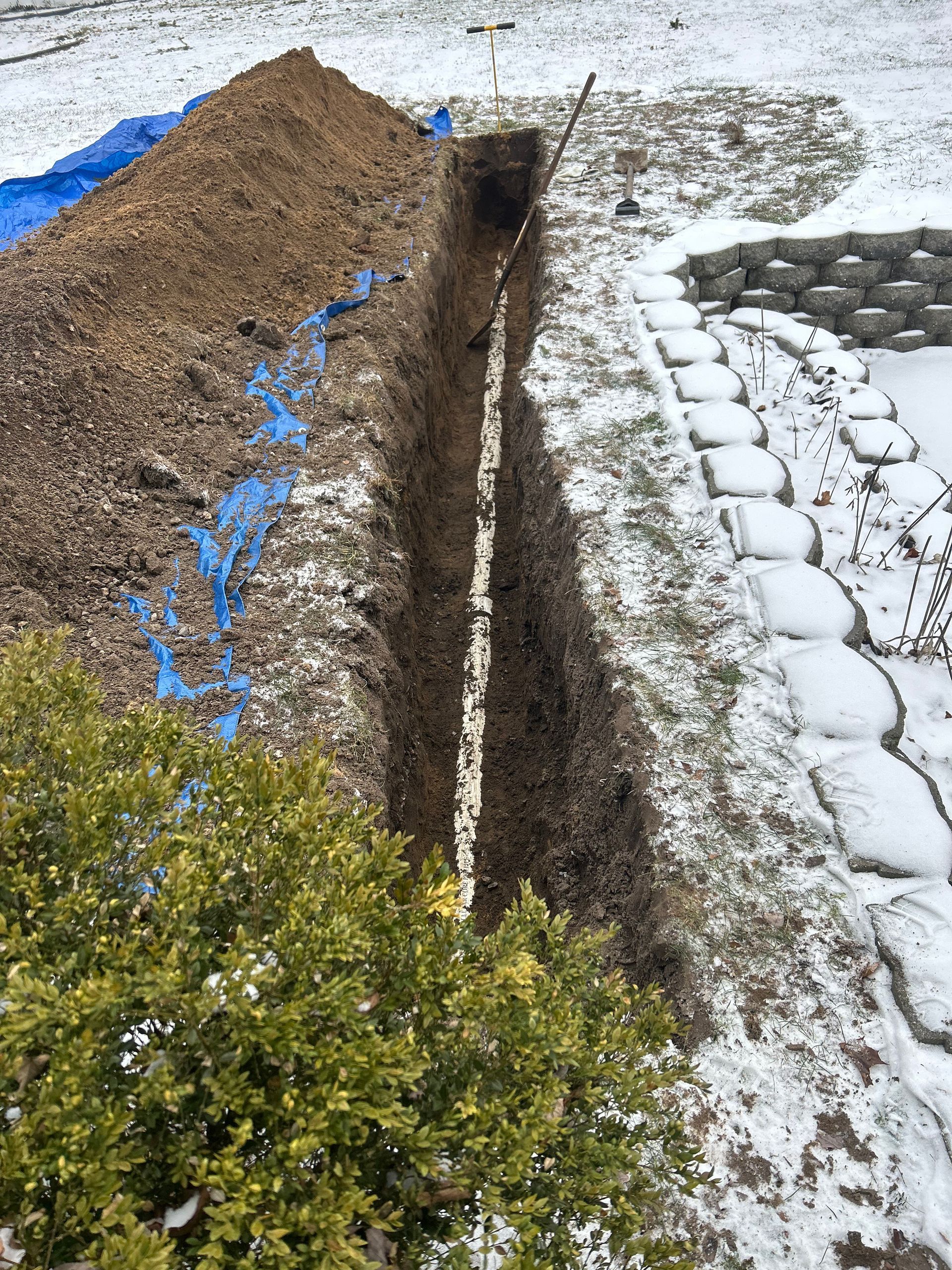 A trench is being dug in the snow next to a pile of dirt.
